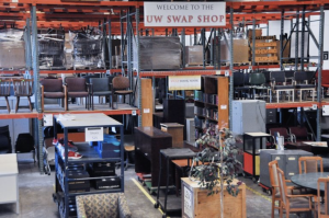 The inside of a warehouse filled with a variety of furniture and other items such as chairs, books, tables, and file cabinets. A sign is hung above materials reading "Welcome to the UW Swap Shop" 